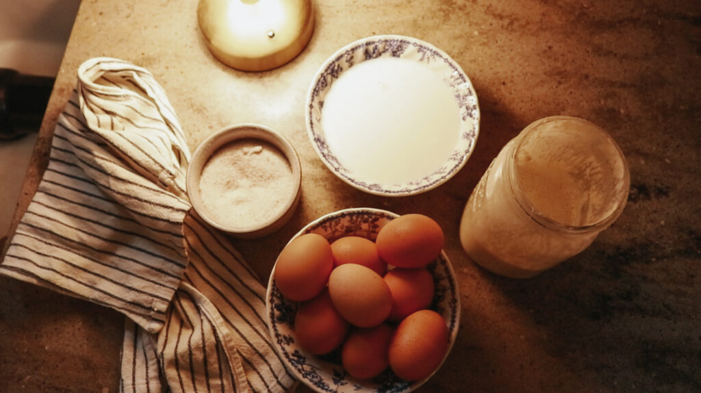 Eggs, sourdough starter, salt, and milk sit on the counter in the glow of a small lamp.