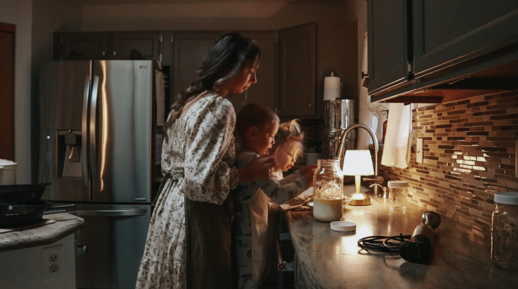 A Christian homemaker in a floral dress and green apron leans over to watch as her two children help in the kitchen.
