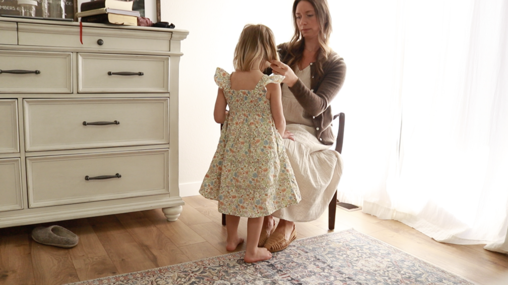 A Christian homemaker in a tan gingham dress fixes her daughter hair. Her daughter is in a floral dress.