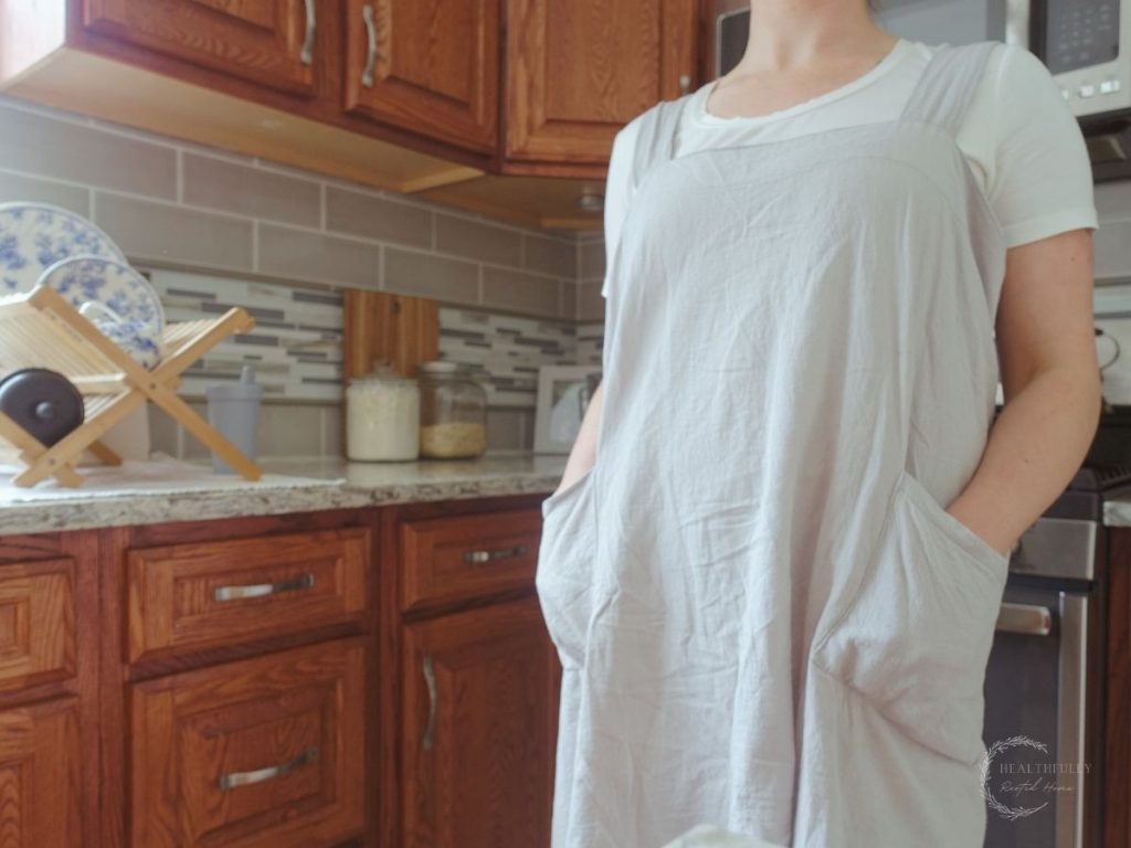 woman wearing a pinafore apron with her hands in the pockets with a drying rack full of dishes in the background