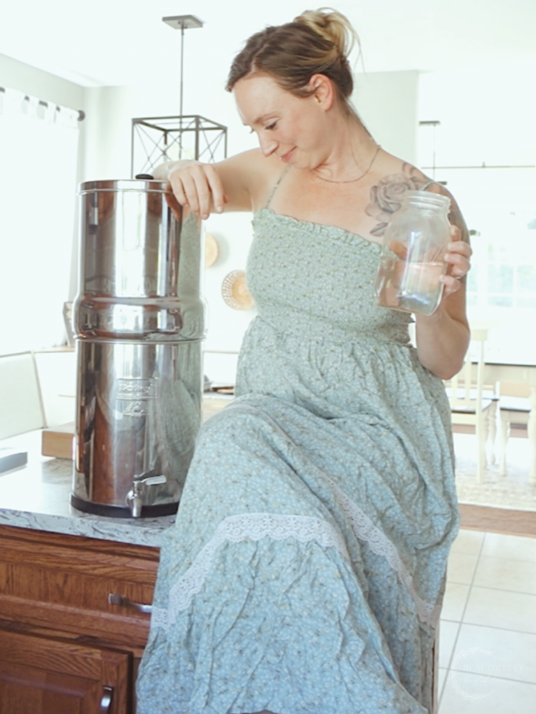 kyrie with healthfully rooted home sitting next to her berkey water filter sitting on top a kitchen island with a glass of filtered water in hand