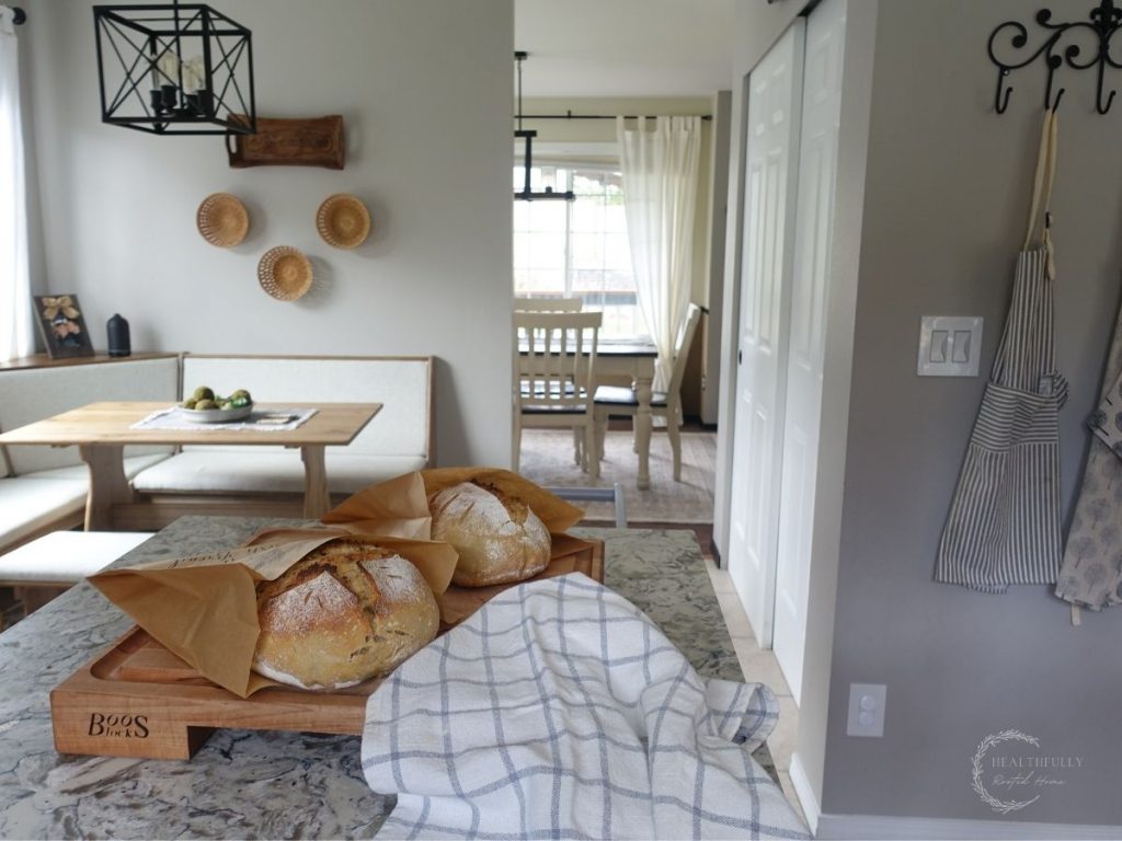 a kitchen with a breakfast nook in the background and two loaves of sourdough bread on the island countertop being stored in brown paper bags