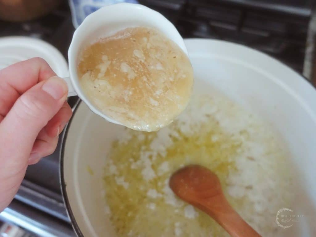 pouring chicken collagen soup into a white dutch oven with onions and olive oil inside