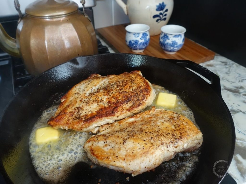 chicken breasts in a cast iron skillet with 2 pads of butter on either side with a copper tea kettle behind it