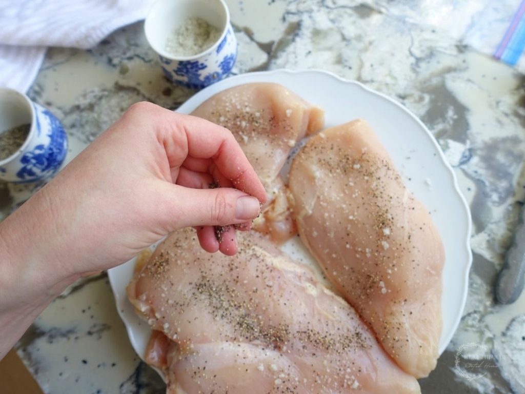 adding slat and pepper to chicken breasts on a white plate with salt and pepper jars on the side