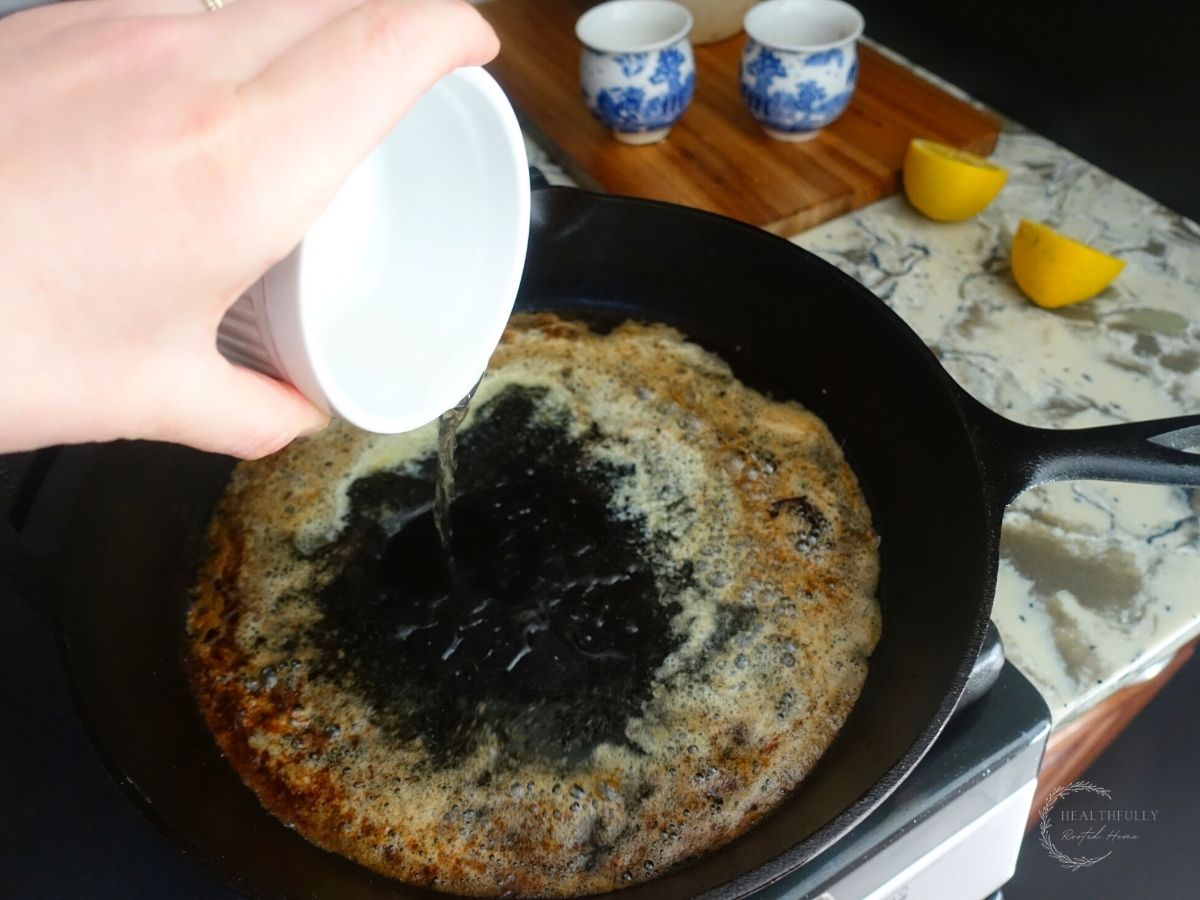 adding white wine vinegar to a cast iron skillet to deglaze the pan with lemon wedges behind it