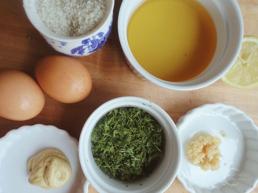 sea salt, avocado oil, brown eggs, fresh chopped dill, garlic and dijon mustard all in white containers on a wooden cutting board