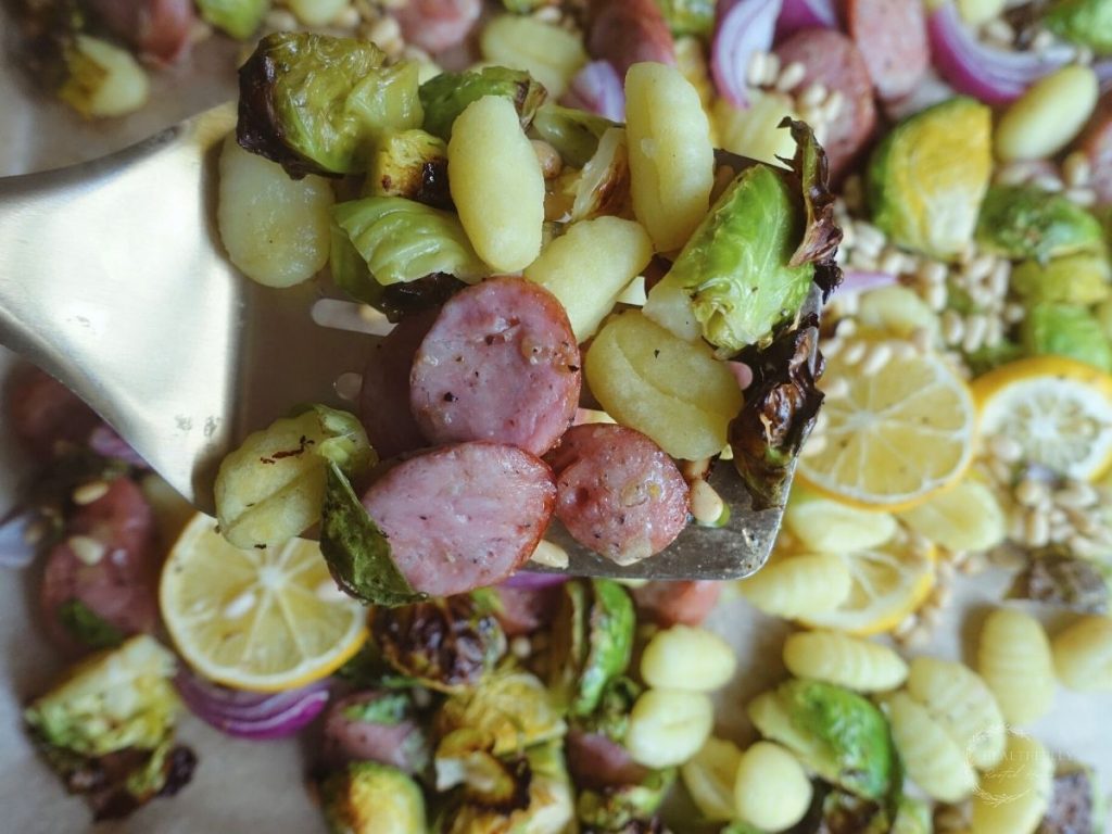 gnocchi tray bake on a gold spatula with chicken apple sausage brussels sprouts red onion pine nuts