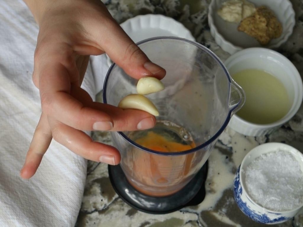 adding garlic cloves to the cup of an immersion blender with other ingredients surrounding it