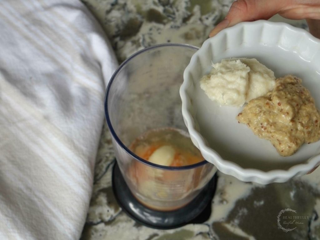adding dijon mustard and horseradish into the cup of an immersion blender with a tea towel to the side