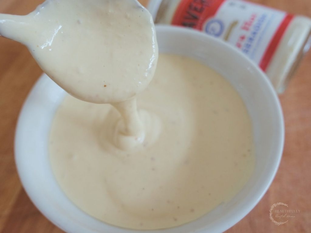 drizzling horseradish aioli from a spoon into a bowl of the sauce with a jar of beaver prepared horseradish in the background