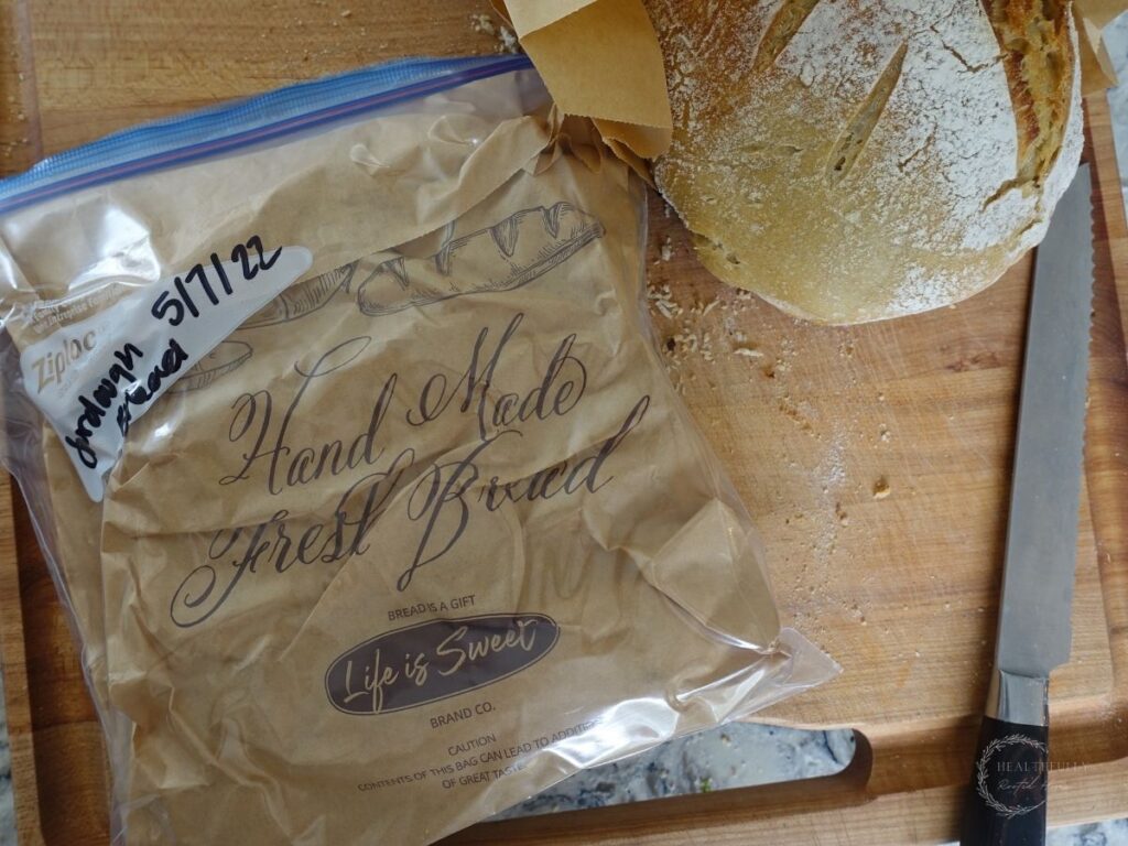 sourdough bread being stored in a plastic bag next to a loaf being stored in a brown paper bag on a wooden cutting board with a bread knife next to them
