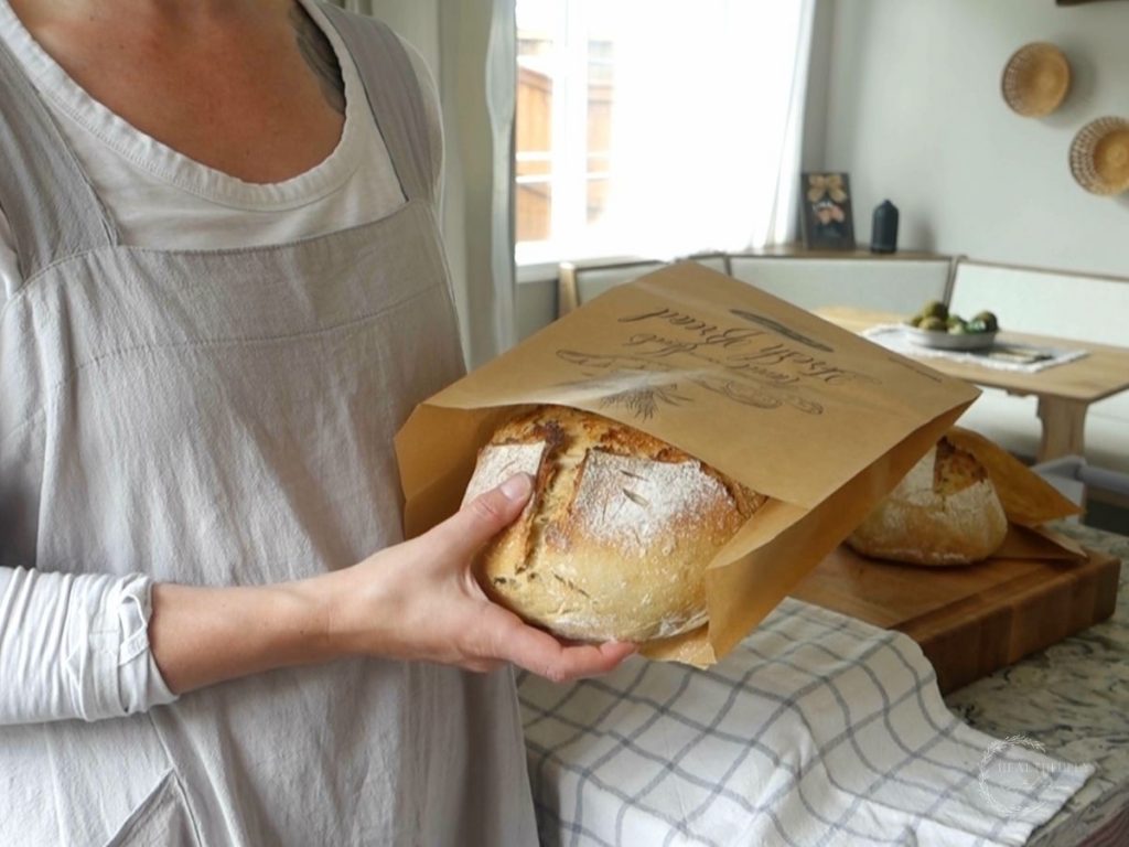 kyrie with healthfully rooted home wearing a pinafore apron placing a loaf of sourdough bread in a brown paper bag for storage