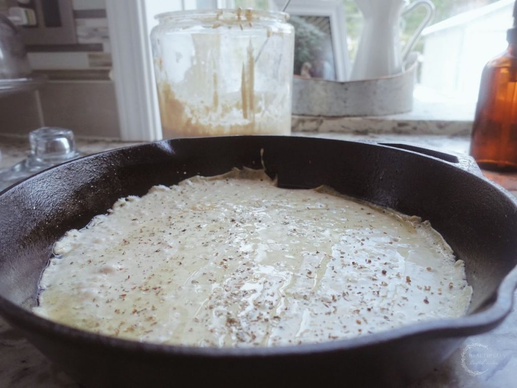 sourdough starter spread around the entire surface of a cast iron skillet with italian seasoning and a jar of sourdough starter in the background