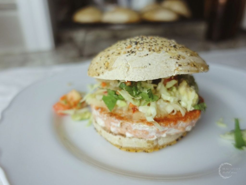 salmon burger on a white plate with sourdough burger buns on a wire cooling rack behind it