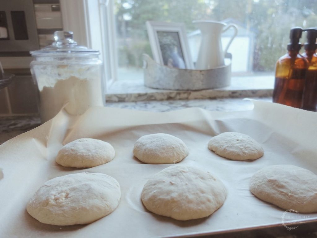 lwtting sourdough burger bun dough proof at room temperature on a parchment line baking sheet with a glass jar of flour in the background