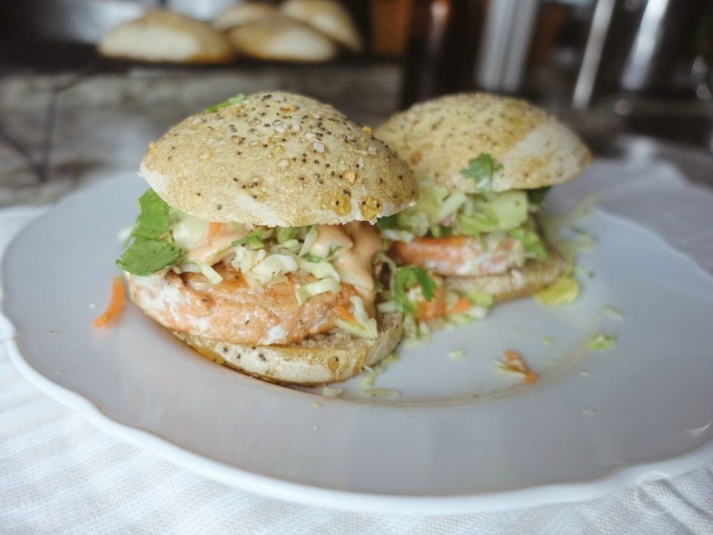 two salmon burgers next to each other on a white plate with more burger buns in the background on a cooling rack 