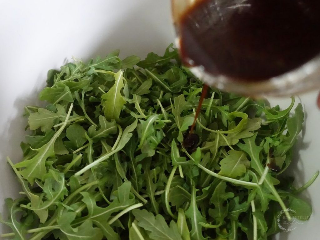 drizzling balsamic vinaigrette on arugula in a white bowl