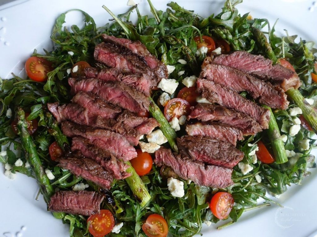 steak and arugula salad with grilled ribeye, grilled asparagus, balsamic dressed arugula cherry tomatoes and smoked blue cheese on a white platter