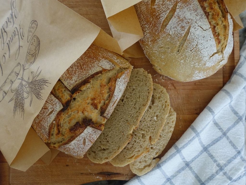 two loaves of sourdough bread being stored in a brown paper bag one loaf is sliced and one isnt