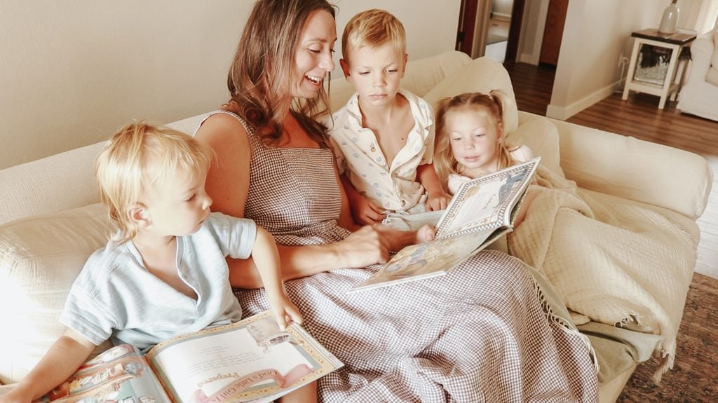 Christian mother sits on the couch reading storybooks aloud with her three children gathered around her.