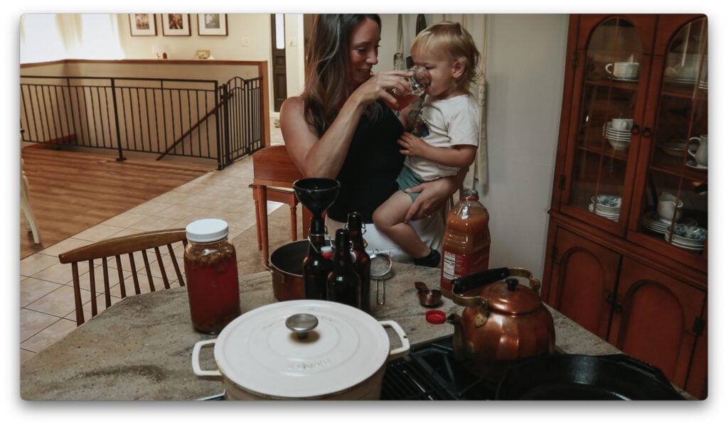 Christian homemaker wearing a black tank top gives her toddler a drink of apple cider while standing in the kitchen.