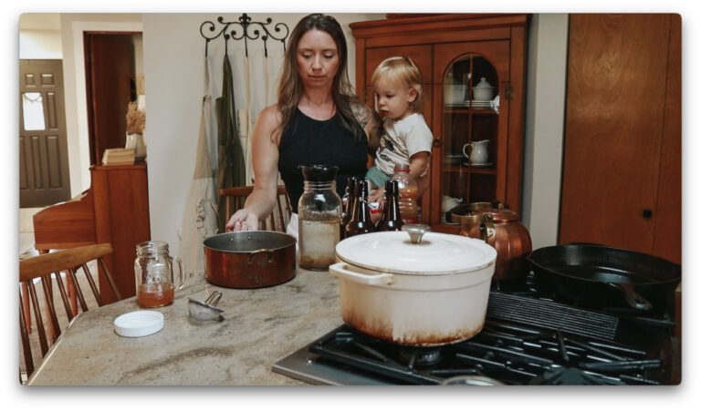 Christian homemaker wearing a black tank top gives her toddler a drink of apple cider while standing in the kitchen.