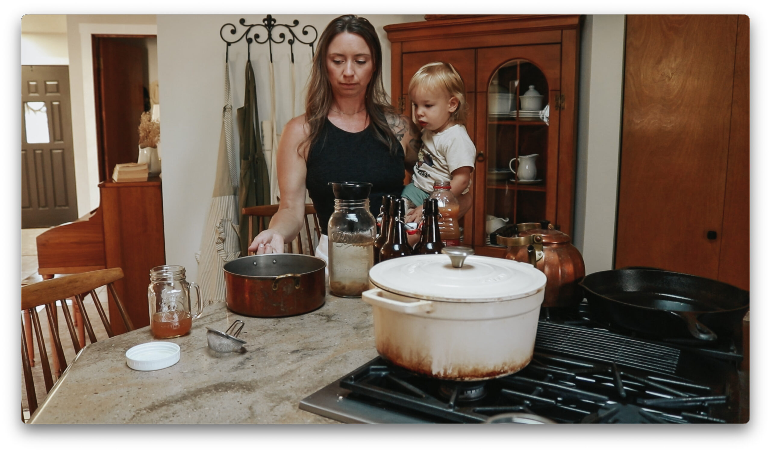 Christian homemaker wearing a black tank top gives her toddler a drink of apple cider while standing in the kitchen.